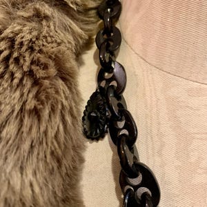 May include: A close-up of a black chain necklace with a decorative pendant. The necklace is made of large, oval-shaped links. The pendant is a dark, ornate design. A brown fur collar is visible on the left side of the image.