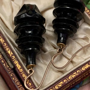 May include: A pair of black, carved earrings with a tiered design. The earrings have gold-colored hooks and are displayed in a decorative box with a cream-colored interior and ornate gold trim. The earrings have a vintage aesthetic.