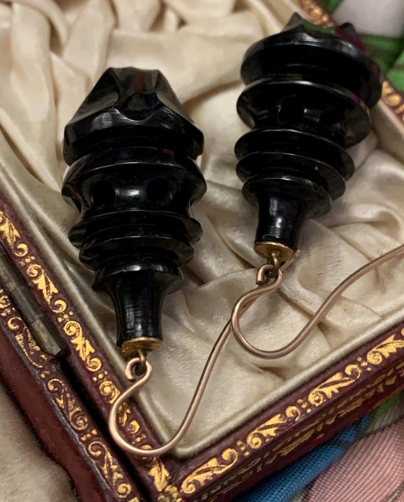 May include: A pair of black, carved earrings with a tiered design. The earrings have gold-colored hooks and are displayed in a decorative box with a cream-colored interior and ornate gold trim. The earrings have a vintage aesthetic.