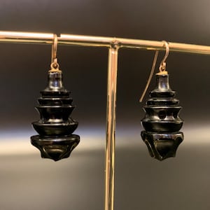 May include: A pair of black, tiered drop earrings with gold-colored ear wires. The earrings have a pagoda-like design, with multiple levels decreasing in size. The earrings are displayed on a gold-colored earring stand against a dark background.