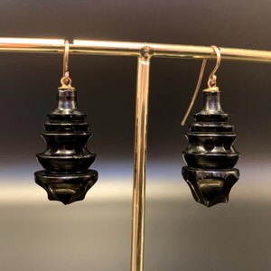 May include: A pair of black, pagoda-shaped earrings with gold-colored ear wires. The earrings have a tiered design, with each level slightly smaller than the one below. The earrings are displayed on a gold-colored earring stand.