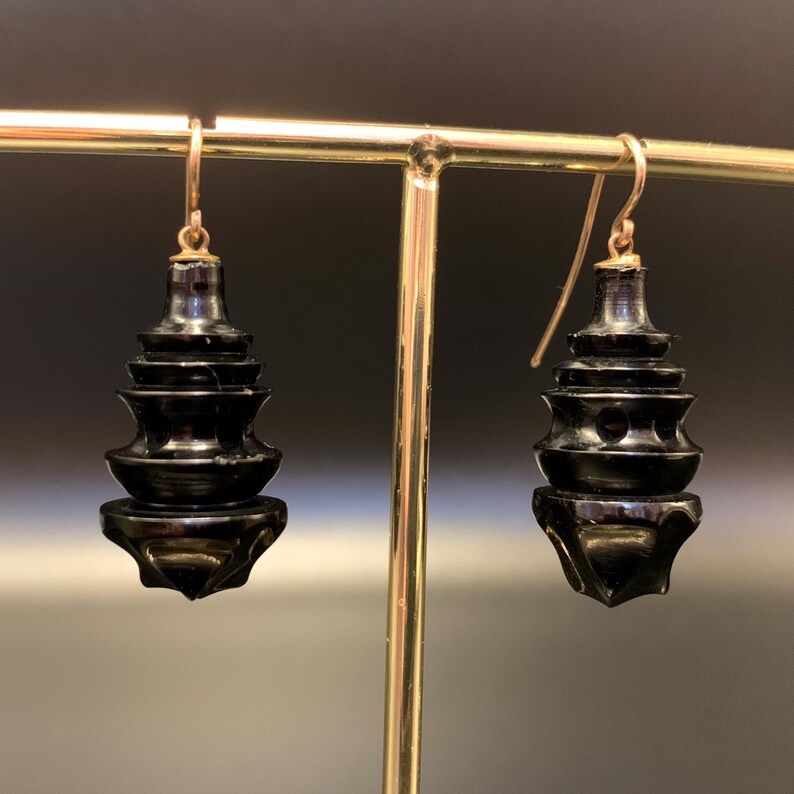 May include: A pair of black, pagoda-shaped earrings with gold-colored ear wires. The earrings have a tiered design, with each level slightly smaller than the one below. The earrings are displayed on a gold-colored earring stand.
