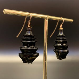 May include: A pair of black, tiered earrings with gold-colored hooks. The earrings have a pagoda-like design, with multiple layers decreasing in size towards the top. The bottom of each earring has a decorative, geometric shape.