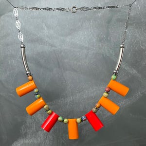 Puede incluir: Un collar hecho a mano con una cadena plateada y una serie de cuentas cilíndricas de colores. El collar presenta cuentas de color naranja y rojo vibrantes, intercaladas con cuentas más pequeñas y multicolores. El diseño se completa con detalles plateados.