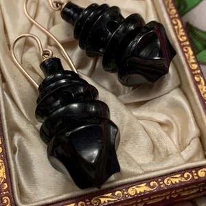 May include: A pair of black, carved earrings with gold-colored ear wires, displayed in a vintage jewelry box. The earrings have a spiral design with a geometric base. The box has a cream-colored satin lining and a decorative, ornate frame.