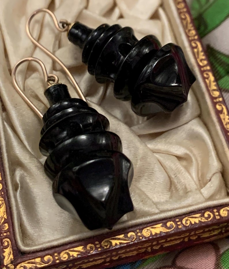 May include: A pair of black, carved earrings with gold-colored ear wires, displayed in a vintage jewelry box. The earrings have a spiral design with a geometric base. The box has a cream-colored satin lining and a decorative, ornate frame.