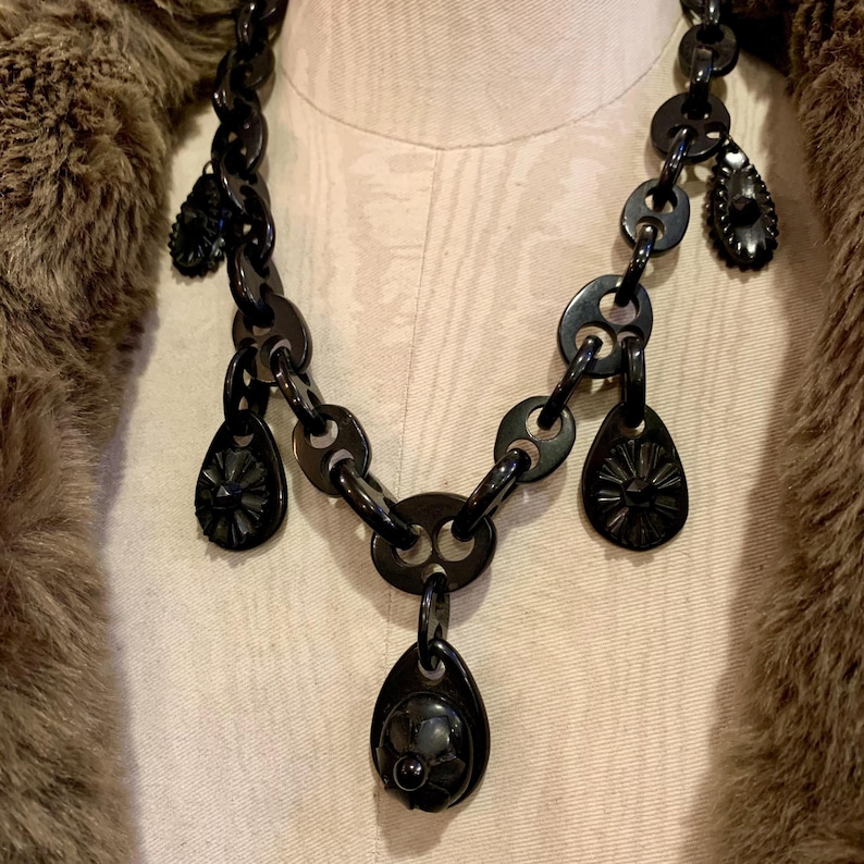 May include: A black statement necklace with a chunky chain design. The necklace features teardrop and circular pendants with intricate floral and geometric patterns. The jewelry is displayed on a mannequin, with a brown fur-like garment in the background.