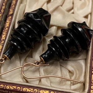 May include: A pair of black, carved earrings with a spiral design. The earrings have gold-colored ear wires and are displayed in a vintage jewelry box with a cream-colored satin lining.
