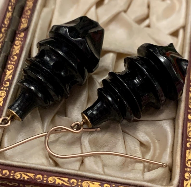 May include: A pair of black, carved earrings with a spiral design. The earrings have gold-colored ear wires and are displayed in a vintage jewelry box with a cream-colored satin lining.