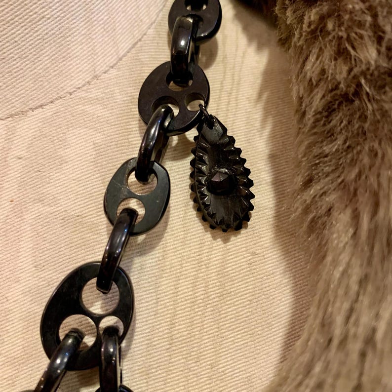 May include: A close-up of a black necklace with large, oval-shaped links. A decorative, dark-colored pendant hangs from the chain. The necklace is displayed against a neutral background, with a portion of a fur collar visible.