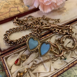 May include: An assortment of antique gold jewellery, featuring a chain bracelet, a heart-shaped locket with a light blue interior, and a pendant with a red gemstone. The jewellery is presented in a vintage box with a map background.