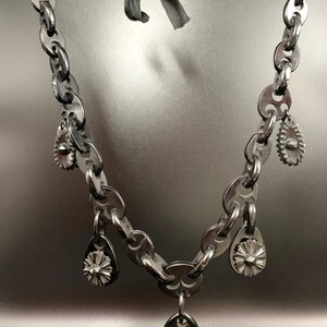 May include: A silver-toned necklace featuring a chain-link design with multiple teardrop-shaped charms and a central pendant. The necklace is suspended by a gray ribbon, creating a unique and edgy aesthetic. The charms have a floral design.