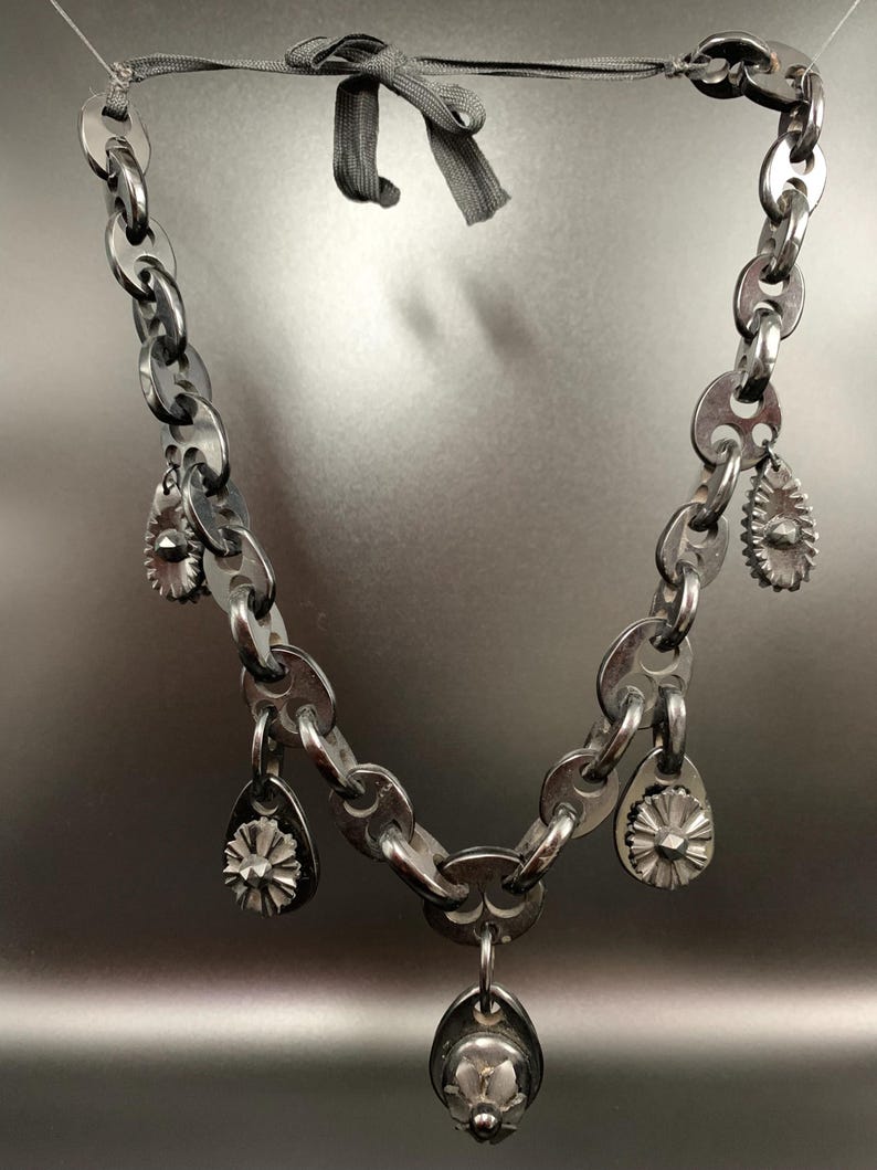 May include: A silver-toned necklace featuring a chain-link design with multiple teardrop-shaped charms and a central pendant. The necklace is suspended by a gray ribbon, creating a unique and edgy aesthetic. The charms have a floral design.