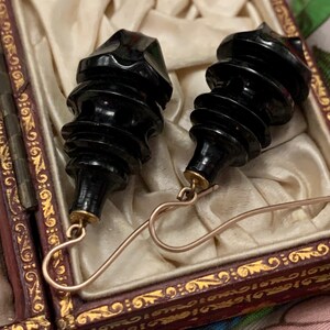 May include: A pair of vintage black earrings with a unique, carved design. The earrings have a tiered, cylindrical shape with a faceted top and are suspended from gold-colored ear wires. They are presented in a decorative, ornate box with a cream-colored satin lining.