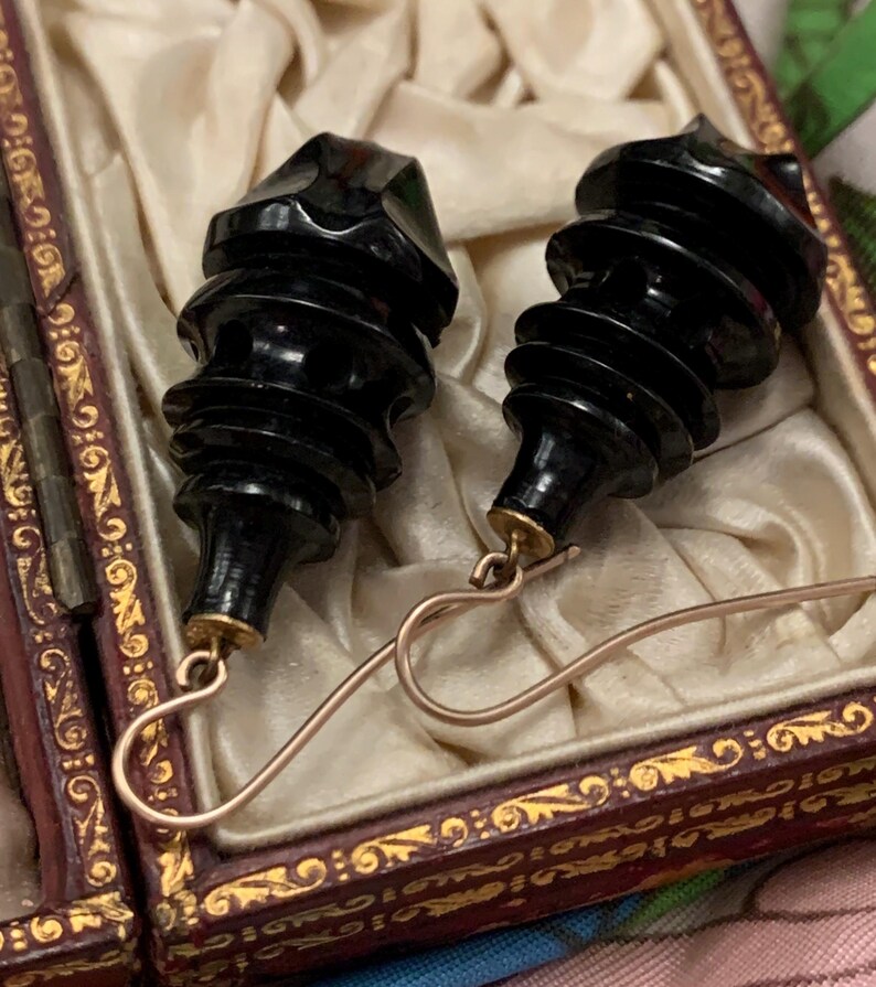 May include: A pair of vintage black earrings with a unique, carved design. The earrings have a tiered, cylindrical shape with a faceted top and are suspended from gold-colored ear wires. They are presented in a decorative, ornate box with a cream-colored satin lining.