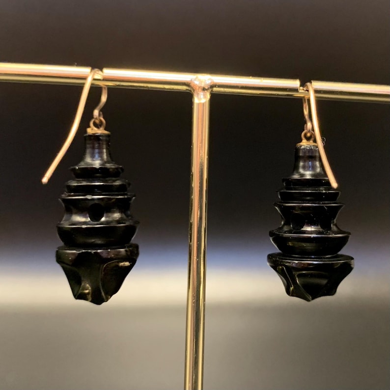 May include: A pair of black, pagoda-shaped earrings with gold-colored ear wires. The earrings have a tiered design, with each tier decreasing in size towards the top. The earrings are displayed on a gold-colored stand against a dark background.
