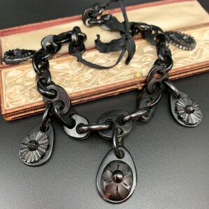 May include: A black vintage necklace with a chain-link design and decorative floral pendants. The necklace is displayed in an open, antique jewelry box with a cream-colored satin lining. The necklace is made of a dark, possibly carved material.