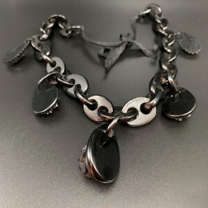 May include: A silver-toned bracelet with a chain-link design and black accents. The bracelet features several small, heart-shaped charms and a larger, teardrop-shaped pendant. The charms have a textured appearance, and the bracelet is displayed on a dark surface.