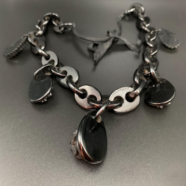 May include: A silver-toned bracelet with a chain-link design and black accents. The bracelet features several small, heart-shaped charms and a larger, teardrop-shaped pendant. The charms have a textured appearance, and the bracelet is displayed on a dark surface.