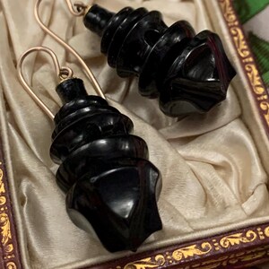 May include: A pair of black, spiral-shaped earrings with gold-colored ear wires. The earrings are displayed in a vintage, ornate box with a cream-colored satin lining. The box has a dark red frame with gold detailing.