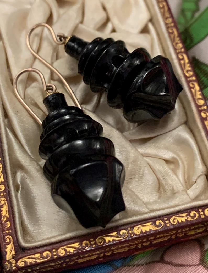 May include: A pair of black, spiral-shaped earrings with gold-colored ear wires. The earrings are displayed in a vintage, ornate box with a cream-colored satin lining. The box has a dark red frame with gold detailing.
