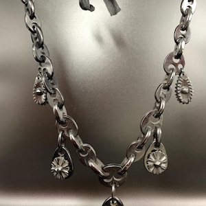 May include: A silver-toned necklace with a chain-link design. The necklace features several teardrop-shaped charms and a central pendant, all with floral-like details. A gray ribbon is used to fasten the necklace.