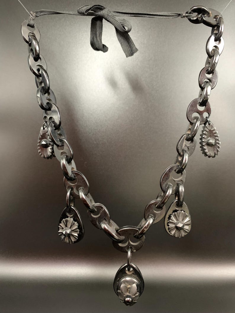 May include: A silver-toned necklace with a chain-link design. The necklace features several teardrop-shaped charms and a central pendant, all with floral-like details. A gray ribbon is used to fasten the necklace.