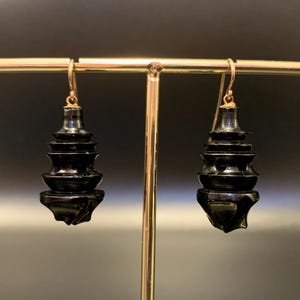 May include: A pair of black, tiered earrings with gold-colored hooks. The earrings have a glossy finish and a unique, stacked design. The earrings are displayed on a gold-colored jewelry stand against a dark background.