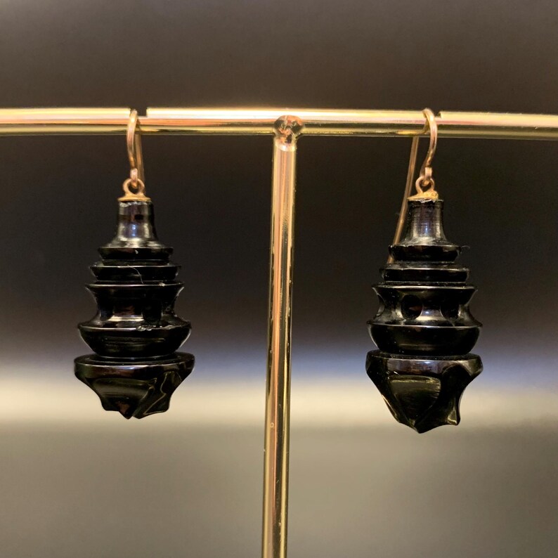 May include: A pair of black, tiered earrings with gold-colored hooks. The earrings have a glossy finish and a unique, stacked design. The earrings are displayed on a gold-colored jewelry stand against a dark background.