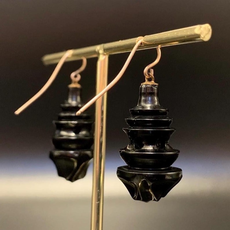 May include: Black, tiered earrings with a pagoda-like design. The earrings are suspended from gold-colored ear wires and are displayed on a gold-colored stand. The earrings have a glossy finish and a unique, layered structure.