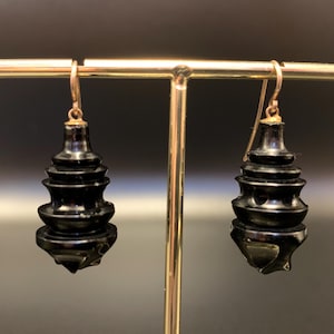 May include: A pair of black, tiered earrings with gold-colored ear wires. The earrings have a unique, pagoda-like design, with multiple stacked levels. The earrings are displayed on a gold-colored stand against a dark background.