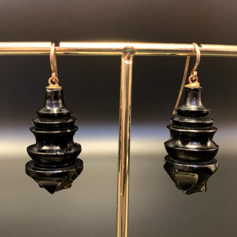 May include: A pair of black, tiered earrings with gold-colored ear wires. The earrings have a unique, pagoda-like design, with multiple stacked levels. The earrings are displayed on a gold-colored stand against a dark background.