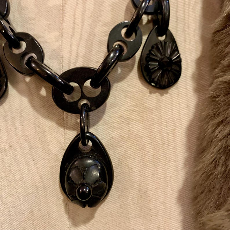 May include: A close-up of a black necklace with a variety of linked shapes and teardrop-shaped pendants. The pendants have a floral design. The necklace is set against a neutral background.