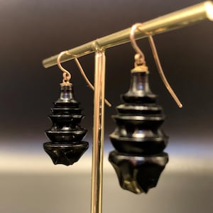 May include: A pair of black, tiered earrings with gold-colored ear wires. The earrings have a pagoda-like design, with multiple levels decreasing in size. The earrings are displayed on a gold-colored earring stand against a dark background.