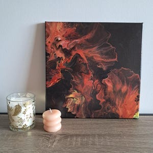 Può includere: Un dipinto astratto con tonalità rosse e arancioni infuocate su sfondo nero. Una piccola candela rosa rotonda e un portacandele in vetro con disegni a foglia d'oro sono davanti all'opera d'arte. Il dipinto è su una tela quadrata.