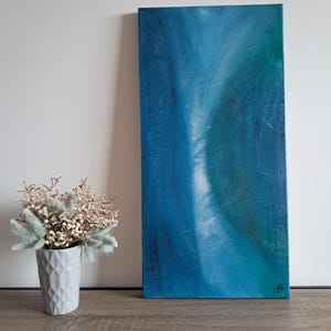 Könnte beinhalten: Abstrakte blaue Leinwandkunst mit strukturierter Oberfläche. Das Kunstwerk zeigt Blautöne und Türkis, wodurch ein fließender, ätherischer Effekt entsteht. Links von der Leinwand steht eine kleine Vase mit Trockenblumen.