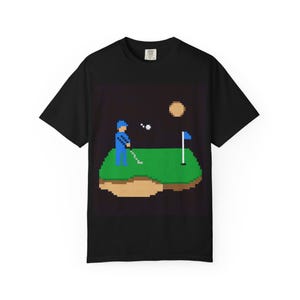 Può includere: T-shirt nero con una scena di golf pixelata. Il design mostra un golfista in blu, un campo da golf verde, una trappola di sabbia, una bandiera e un sole. La pallina da golf è in aria. Una divertente maglietta grafica in stile retrò.