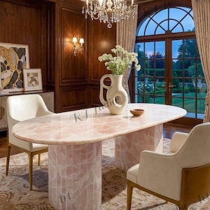 May include: An oval-shaped pink quartz dining table with two cylindrical bases. The table is surrounded by cream-colored chairs and set in a room with dark wood paneling, a chandelier, and a large window overlooking a garden.