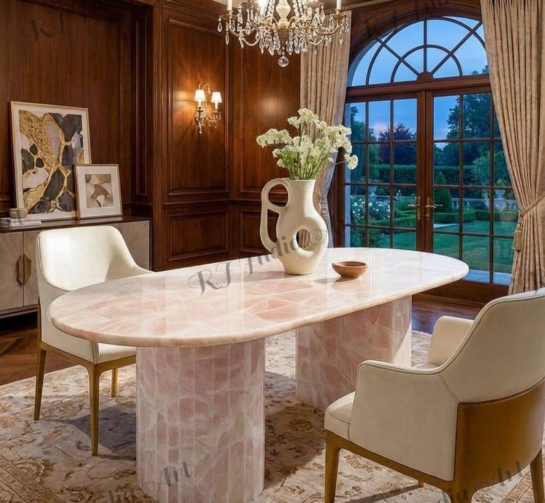 May include: An oval-shaped pink quartz dining table with two cylindrical bases. The table is surrounded by cream-colored chairs and set in a room with dark wood paneling, a chandelier, and a large window overlooking a garden.