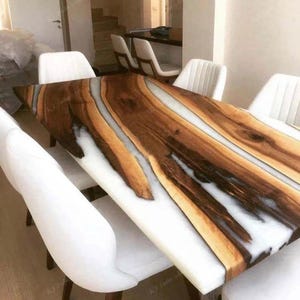 Könnte beinhalten: Ein rechteckiger Esstisch mit einer Tischplatte aus rohem Holz. Das Holz ist von satten Brauntönen, mit weißem Harz, das die Lücken ausfüllt. Der Tisch ist von weißen, gepolsterten Stühlen umgeben und bildet eine moderne und elegante Essgruppe.