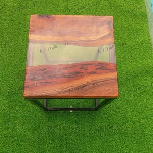 May include: A square wooden side table with a clear resin center. The wood has a rich, reddish-brown colour and is set in a black metal frame. The table is approximately 46 cm square and 46 cm high.