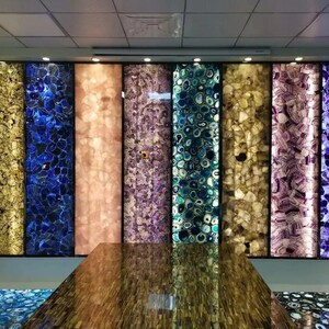 May include: A display of backlit gemstone panels in various colors, including gold, blue, peach, purple, teal, and lavender. The panels are arranged vertically, showcasing the natural patterns and textures of the stones. A table with a reflective surface is in the foreground.