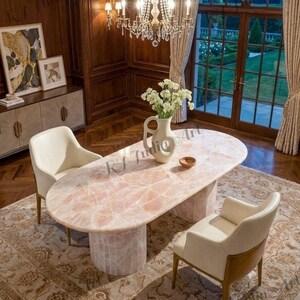 May include: An oval-shaped, pink quartz dining table with a cylindrical base. Two cream-colored chairs are positioned on either side of the table. A vase of white flowers sits on the table. The room has dark wood paneling and a large window.