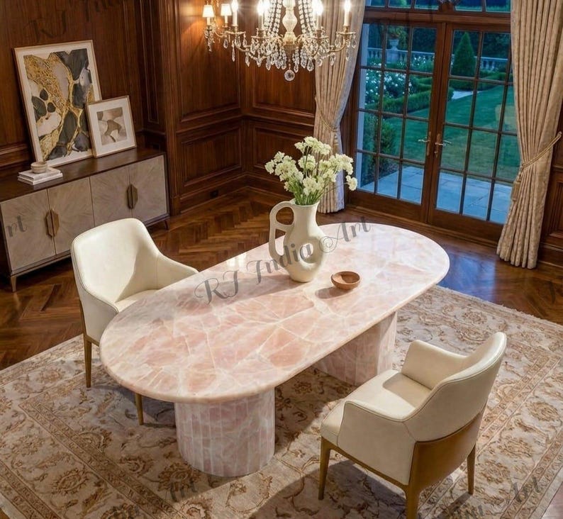 May include: An oval-shaped, pink quartz dining table with a cylindrical base. Two cream-colored chairs are positioned on either side of the table. A vase of white flowers sits on the table. The room has dark wood paneling and a large window.