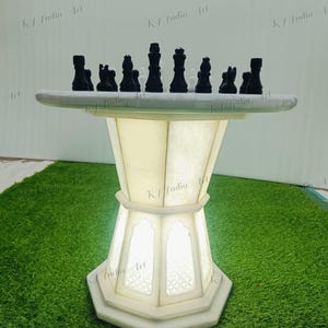 May include: A white marble chess table with black chess pieces. The table has a round top and a geometric base with a light inside. The chess pieces are arranged on the table, ready for a game. The table is on a green surface.