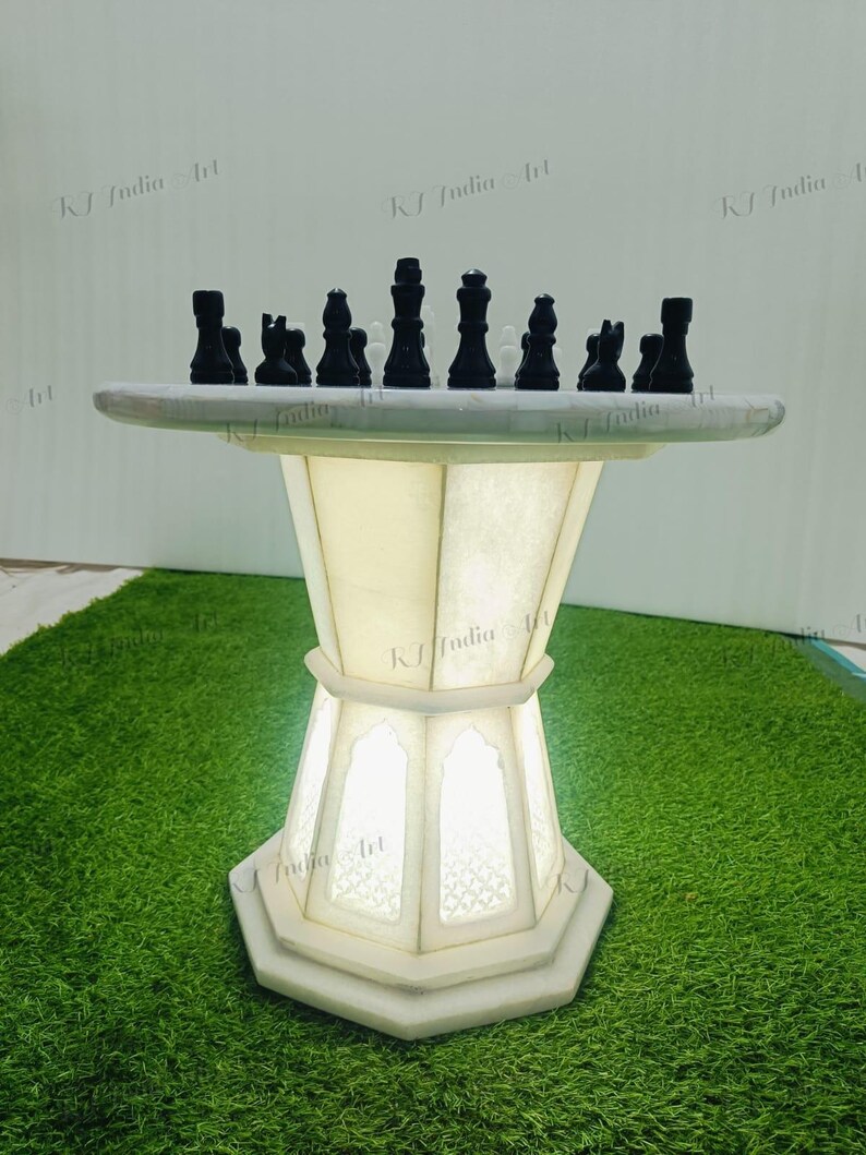 May include: A white marble chess table with black chess pieces. The table has a round top and a geometric base with a light inside. The chess pieces are arranged on the table, ready for a game. The table is on a green surface.