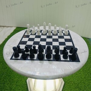 May include: A round chess table with a black and white checkered playing surface. The chess pieces are black and white. The table has a white base with a geometric design. The table is on a green surface.