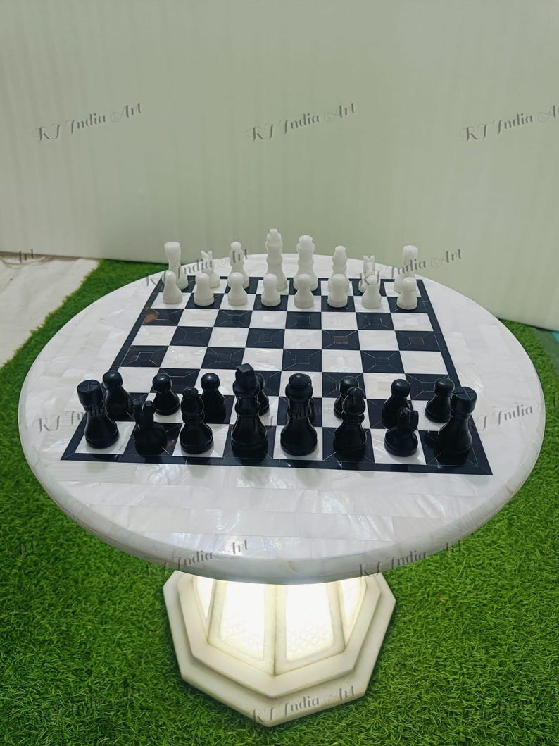 May include: A round chess table with a black and white checkered playing surface. The chess pieces are black and white. The table has a white base with a geometric design. The table is on a green surface.