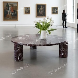 Può includere: Un tavolino rotondo in marmo bordeaux con un vaso di tulipani bianchi. Il tavolo ha un piano circolare e gambe cilindriche. Il tavolo si trova in una galleria con dipinti.