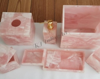 May include: A rose quartz bathroom set including a tissue box, soap dispenser, tray, and other accessories. The set features a light pink color with white veining, creating a natural, elegant look. The soap dispenser has a gold pump.